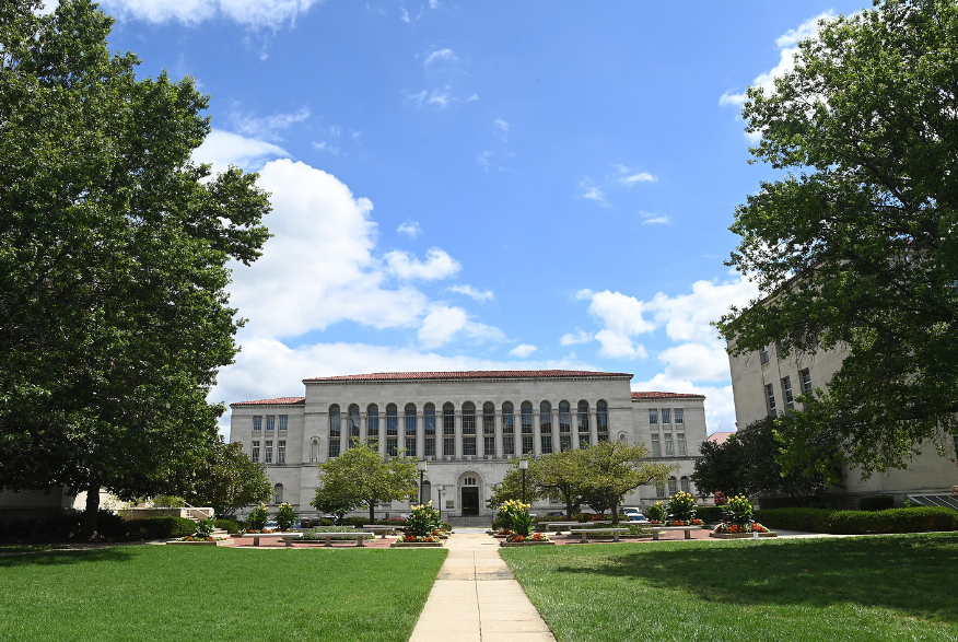 a photo of Mullen Library in the summer: a white building on a large green lawn with a clear blue sky above. 